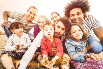 Happy multiracial families taking selfie at beach making funny faces - Multicultural happiness joy and love concept with mixed race people having fun outdoor at sunset - Bright pastel backlight filter