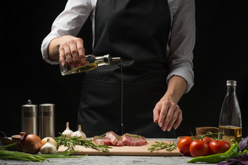 Chef pouring olive oil, three fresh raw steaks with meat on a dark background. Watering with oil before cooking, on a background with vegetables, tomatoes, hot pepper, onions, rosemary and basil