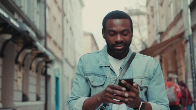 young african amerian man goes down the street and uses phone look at smartwatch smile happy internet face technology fashion black call texting attractive smartphone communication handsome message