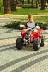 Boy riding big red toy car in sunlit summer park.