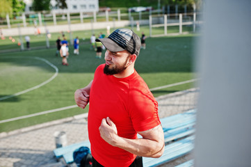 Young brutal bearded muscular man wear on red shirt, shorts and cap at stadium.