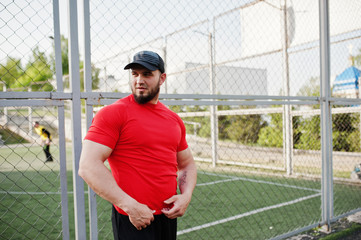 Young brutal bearded muscular man wear on red shirt, shorts and cap at stadium.