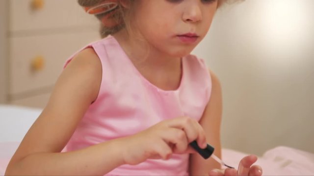 Pretty girl in curlers doing manicure. Little princess in pink dress painting nails sitting on bed. 