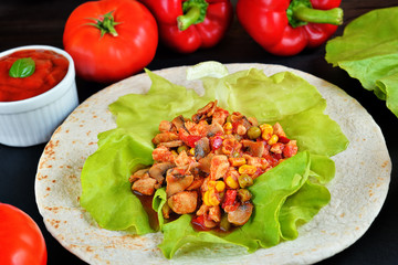 Photo of freshly cooked tasty tortilla wrap with chicken breast and vegetables: pepper, corn, lettuce, tomatoes and champignons on the wooden dark table. Top view on black. Mexican food