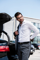 low angle view of handsome businessman taking off jacket and looking away near car