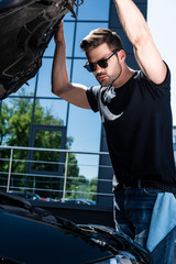 low angle view of young man in sunglasses checking car engine at street