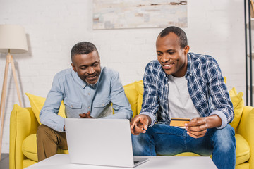 smiling senior man and adult son with credit card using laptop together at home