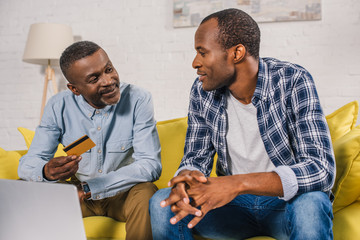 senior man with credit card and adult son smiling each other while using laptop together at home