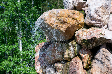 A small waterfall sprinkles drops of water down from a large stone in the form of a rock against the background of green trees. Decorative designer fountain.