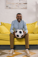 smiling african american man sitting on sofa and holding soccer ball