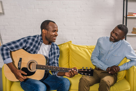 Smiling Senior Man Looking At Adult Son Playing Guitar At Home