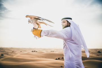 Naklejka premium Arabski mężczyzna w tradycyjnym emirackim stroju spacerujący po pustyni ze swoim sokołem