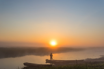 Foggy landscape with a fisherman standing on the river shore at dawn under the rising sun