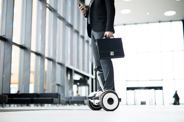 Mobile businessman with briefcase texting in smartphone while moving on hoverboard