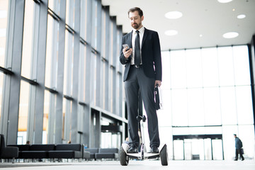 Young berded businessman in suit messaging in smartphone while moving on hoverboard along corridor
