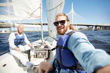Abwaschbare Fototapete Segeln Happy young active man in sunglasses and lifejacket making selfie during sailing with senior friend