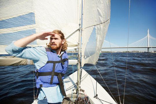 Young bearded man in lifejacket floating on yacht and looking forwards with his hand by forehead