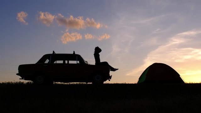 A silhouette of a young woman, sitting on a car and fixing her hair against the sunset