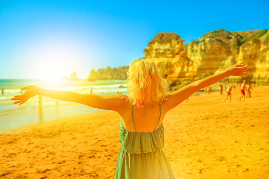 Blonde lifestyle female tourist with raised arms on Praia Dona Ana at sunset. Caucasian woman enjoying at popular Dona Ana Beach in Lagos, Algarve coast. Summer holidays in Europe. Tourism in Portugal