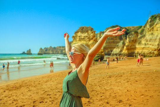 Blonde carefree female tourist with raised arms on Praia Dona Ana in summer holidays. Caucasian woman enjoying at popular Dona Ana Beach in Lagos, Algarve coast. Tourism in Portugal, Europe.