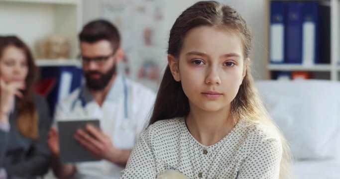 Nice little sick Caucasian patient sitting on hospital bad. Unhappy Ukrainian girl looking attentively at camera on background of mom and doctor. Close-up.