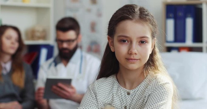 One nice sick girl with sad smile looking directly at camera. Young girl sitting on bad on background of mother and attending physician. Close-up.