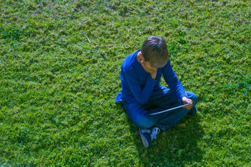 Boy with tablet computer sitting on the grass at sunny day outdoors.