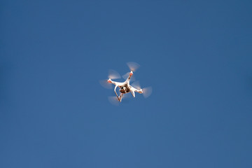 Drone quad copter fly on blue sky in background. Modern drone is flying in air, to take photos and record footage from above. Drone with four propellers, camera and red warning lights. New technology.