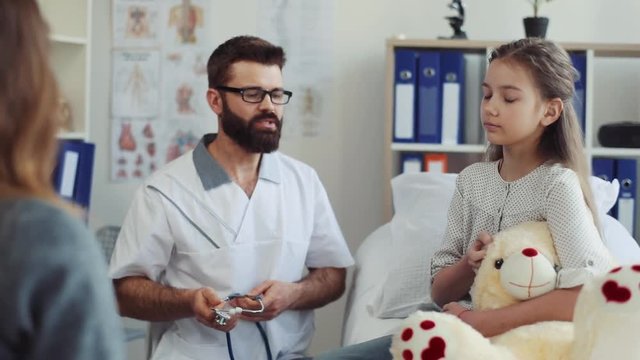 Experienced doctor in glasses listening to little girl, using stethoscope and giving recommendation for recovery. Patient sitting on hospital bad. Indoor. Mother and daughter. Indoors.