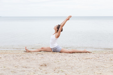 Fototapeta na wymiar A girl with a short hairstyle dressed in shorts and a white jersey makes a twine on the background of the sea and the beach. Playing sports in nature. Stretching, Yoga pose