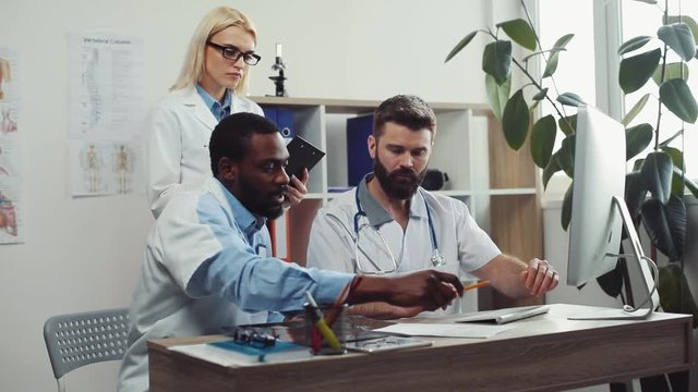 Three multicultural doctors working together in modern doctor&rsquo;s office at prestigious clinic of city. Man with beard typing on keyboard. People looking at medical documents.