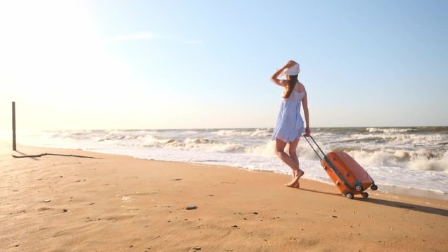 Young woman with a suitcase sitting on the beach. a young girl walks along the beach with a wheeled suitcase.Shags along the sand along the sea. The girl is looking for herself and adventure