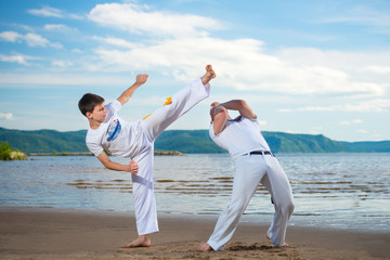 The man is a coach of capoeira and a boy practicing capoeira on the beach. Man and young guy play...