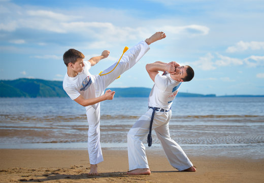 The man is a coach of capoeira and a boy practicing capoeira on the beach. Man and young guy play staged fight.