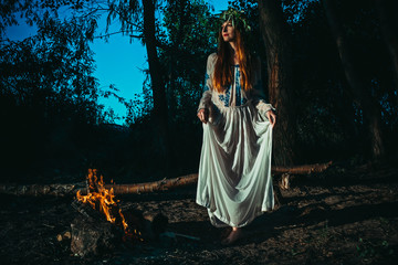 Woman in ethnic clothes with chaplet on a head near bonfire. Concept of Pagan holiday.