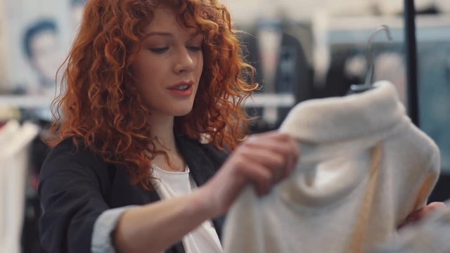 Smiling young woman with curly red hair choose clothes at store hold sweater smile design shopping girl beautiful attractive shop cute happy pretty style curly hair portrait slow motion