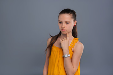 Deep in thought. Petite teenage girl thinking about something intently and touching her jaw with a finger while standing isolated on a blue-grey background