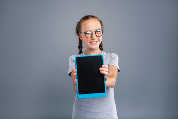 Brand new device. Upbeat teenage girl in glasses posing with a tablet and smiling brightly while standing isolated on a blue-grey background