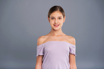 Refined beauty. The portrait of a petite teenage girl in a lilac strapless top smiling at the camera while posing isolated on a grey-blue background