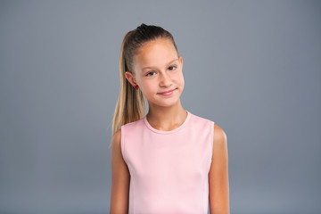 Obraz premium Pretty kid. The portrait of a charming teenage girl with a ponytail smiling at the camera pleasantly and posing isolated on a blue-grey background