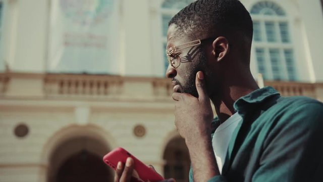 Serious African American young man with glasses using phone stand in city thinking technology businessmen busy people attractive modern black portrait handsome lifestyle smartphone ambitious mobile