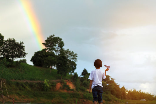 Little Kid Playing Paper Airplane Near Lake And Under Cloudy Sky With Rainbow.