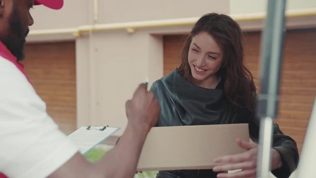 Smiling courier African American gives a parce to a woman after signing on delivery device cardboard technology box order post postal service device sunny beard covered load logistic mailman