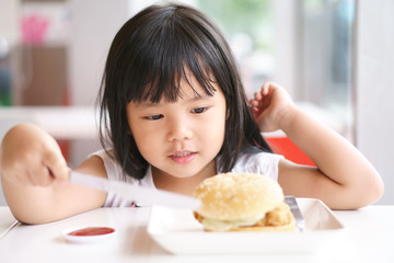 Obraz na płótnie Canvas Asian children cute or kid girl delicious eating ham burger or junk food with tomato sauce or ketchup on white dish and table for breakfast or lunch with hold knife for happy fun smile at restaurant