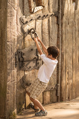 A boy wearing white shirt and checkered shorts pulls the forged door handle of an old textured wooden door.