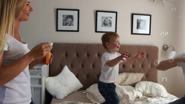 Caring mother plays son looking at soap bubbles, the boy is happy jumping on the bed
