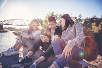 Company school friends teens of different ethnic nationalities sit outside, relax, have fun with smartphone, in the rays of the setting sun