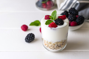 White yogurt with muesli and raspberries in glass bowl on white wooden background.