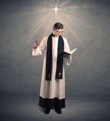 A young male priest in black and white giving his blessing in front of grey wall with glowing cross...