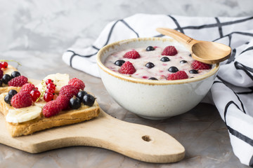 A bowl of oatmeal with berries next to a board with sandwiches with peanut butter and banana, sporodina, raspberries. Healthy summer breakfast on a gray table